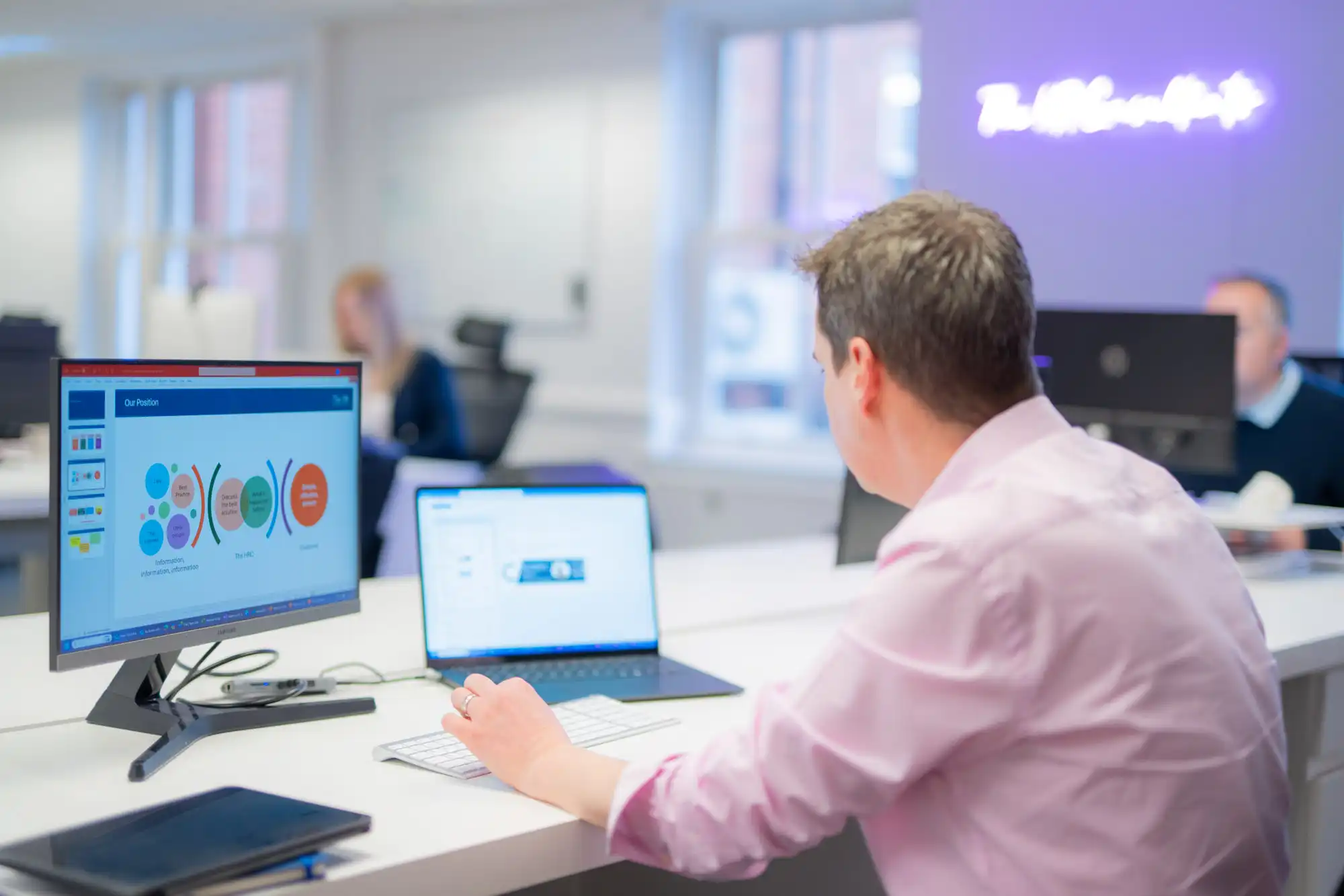 Businessperson working at a desk using a laptop and desktop computer displaying an HR strategy presentation.