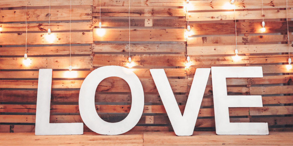Large white LOVE letters against a rustic wood background with warm string lights