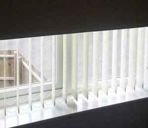 White vertical blinds partially open on a window, showing a fence outside.