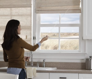 Woman adjusting woven wood blinds in kitchen with beach view. Natural light control.