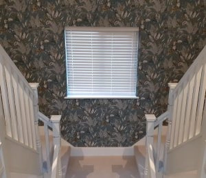Stairwell with white banisters, patterned wallpaper, and a window with white blinds.